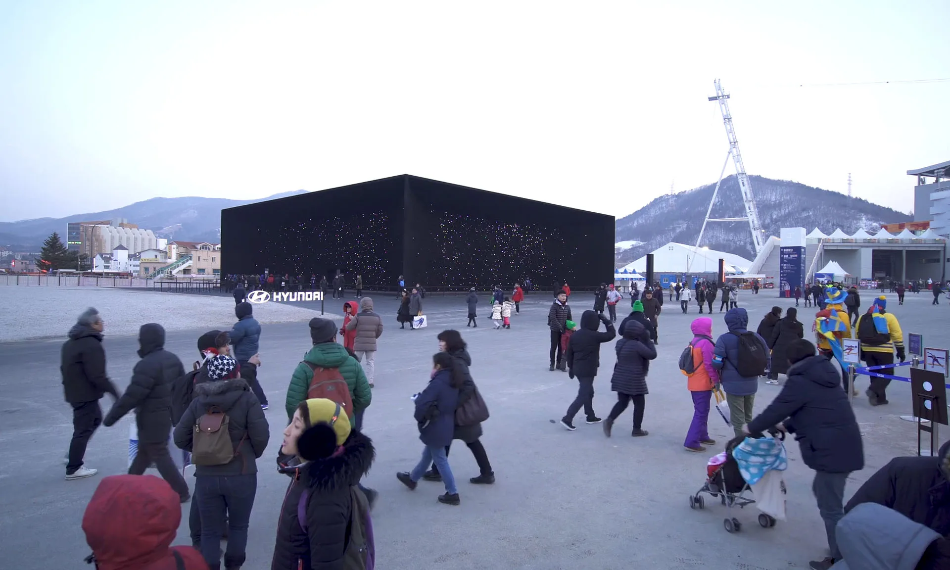 A dense crowd streaming past the pavilion on a bright day, the Hyundai name on the dark wall and the fairground tower beyond
