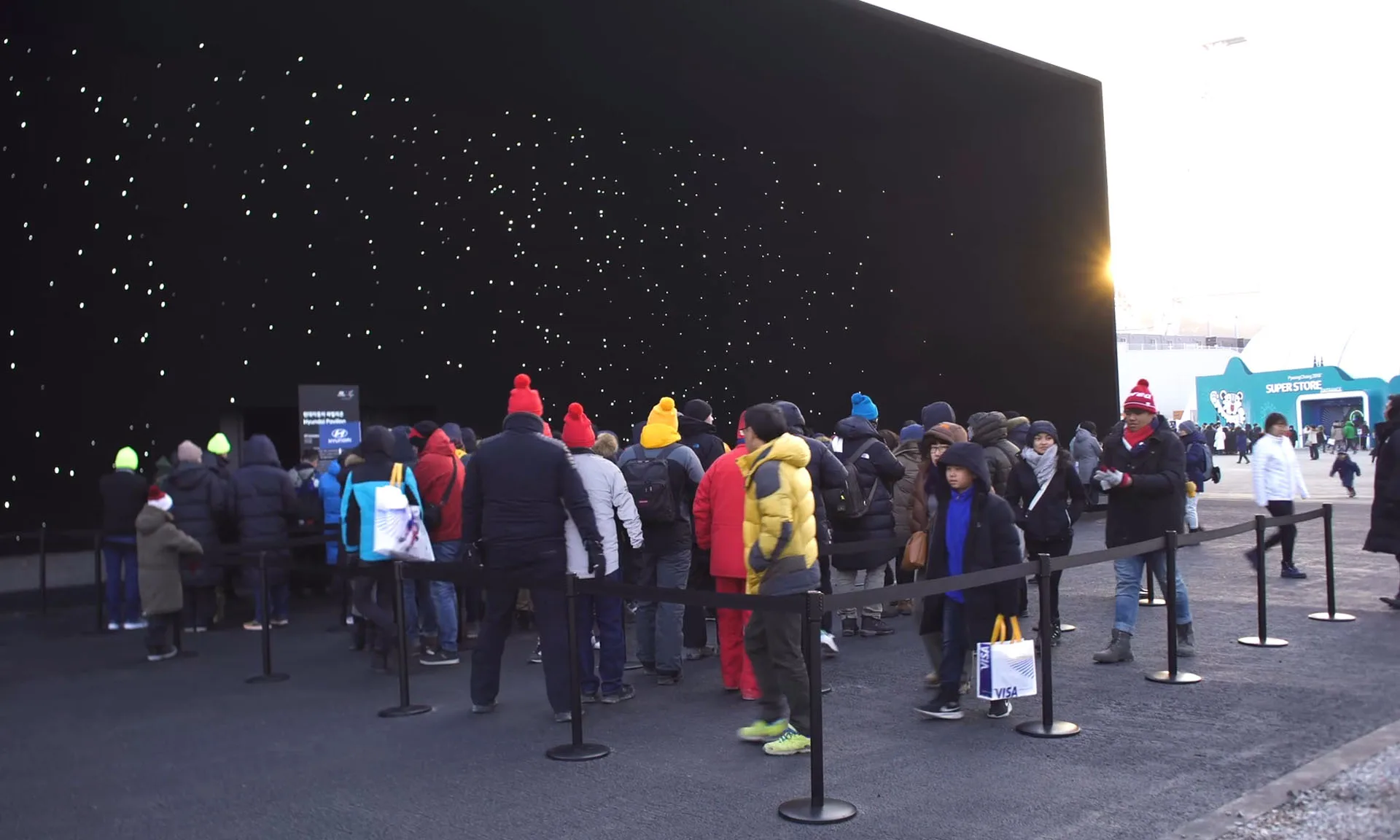 A queue of visitors held behind barrier posts at dusk, the star speckled wall rising black above them