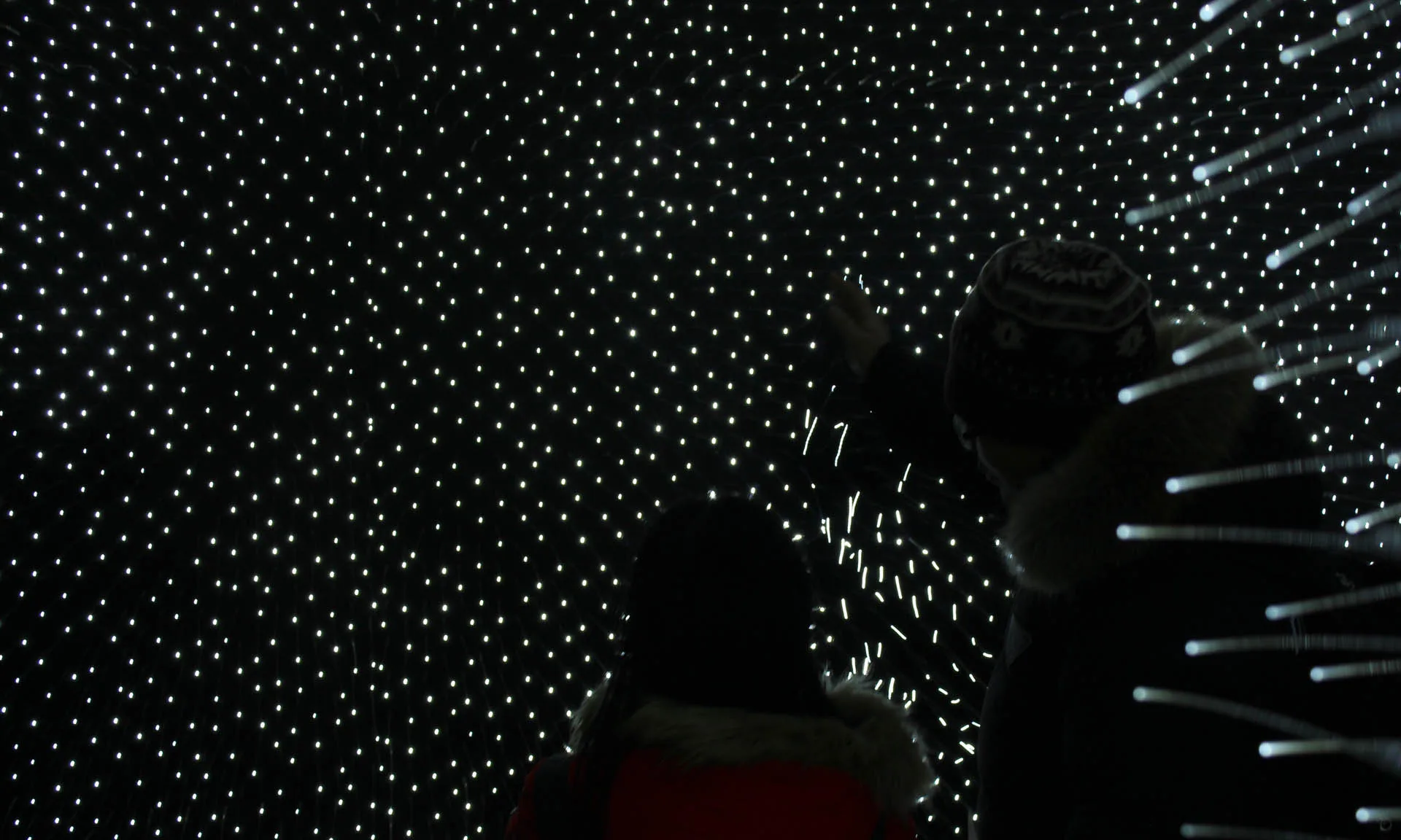Visitors silhouetted against the wall of lights, one of them reaching up with a gloved hand toward the points