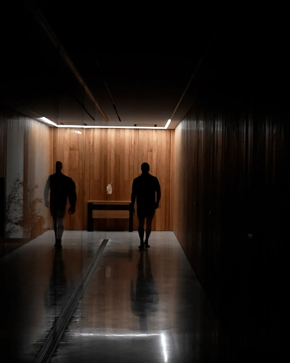 Two people walking away down the dim corridor at night, warm low light on the timber and their reflections in the floor