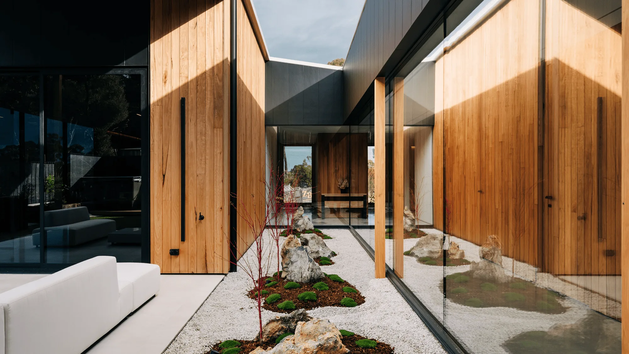 Exterior of the timber-clad pavilions in daylight, a raked gravel and rock courtyard between them and a pool beyond